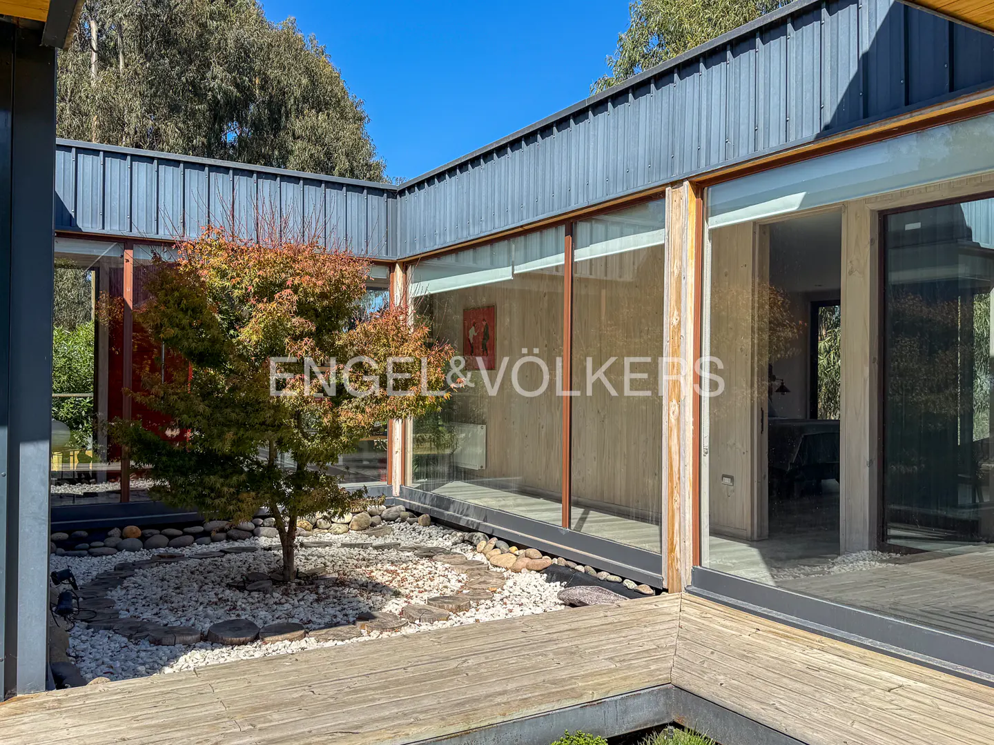 Exterior view of a modern home with a courtyard featuring a small tree, white rocks, and a wooden walkway. Large windows reflect the interior.