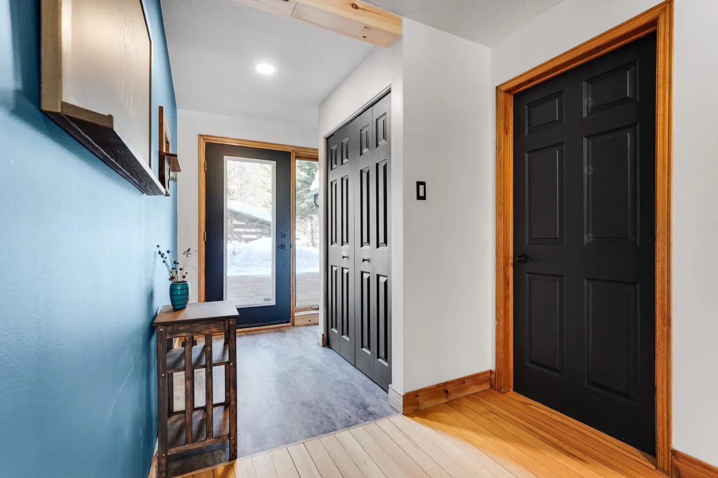 Hallway with blue wall, wood floors, and black doors. A small table with a vase sits near a glass door to a snowy deck.