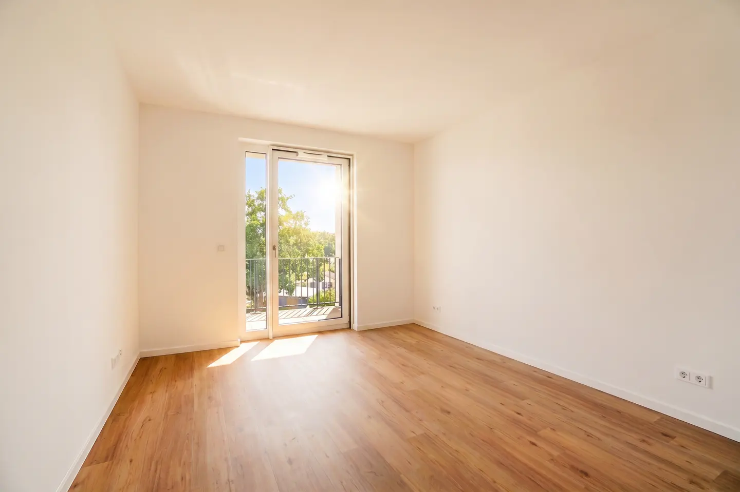 Bright, empty room with wood floors and white walls. Sunlight streams in from a sliding glass door to a balcony.