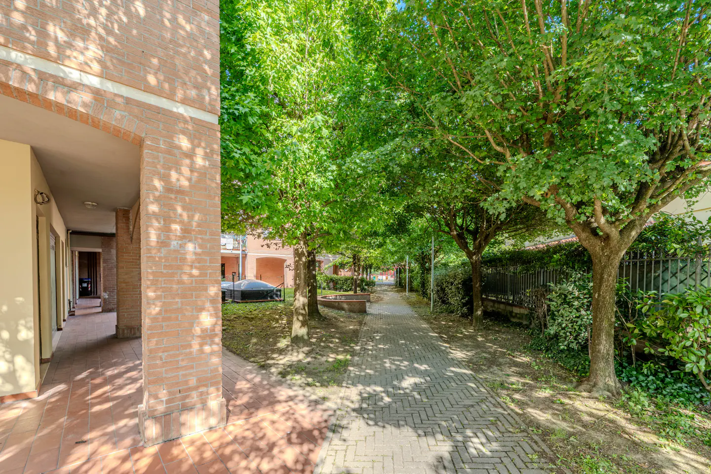 Brick walkway lined with green trees and a brick building with arched openings on the left.