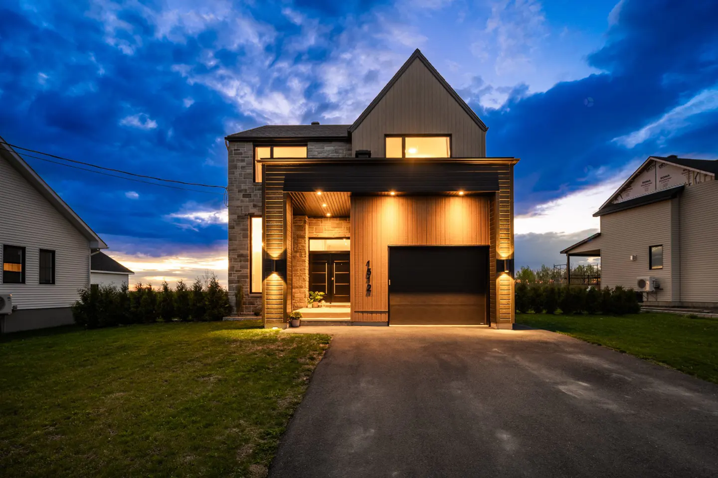 Modern two-story home with stone and wood facade, black garage door, and illuminated exterior lights at dusk.