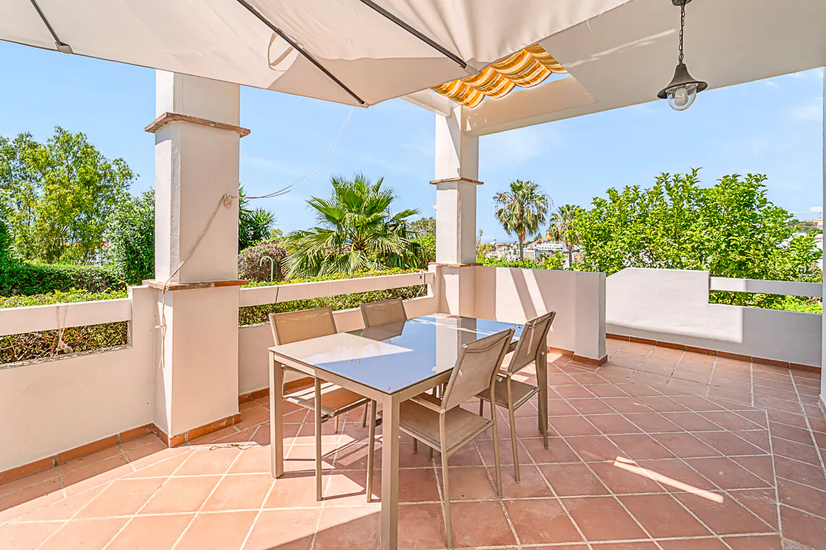 Covered outdoor patio with a glass-top dining table, four chairs, and a scenic view of palm trees and greenery.