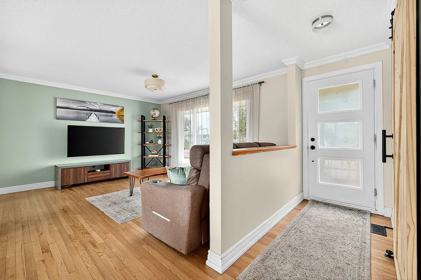 A bright living room and entryway with hardwood floors. A brown recliner faces a TV. A white door with black hardware is on the right.