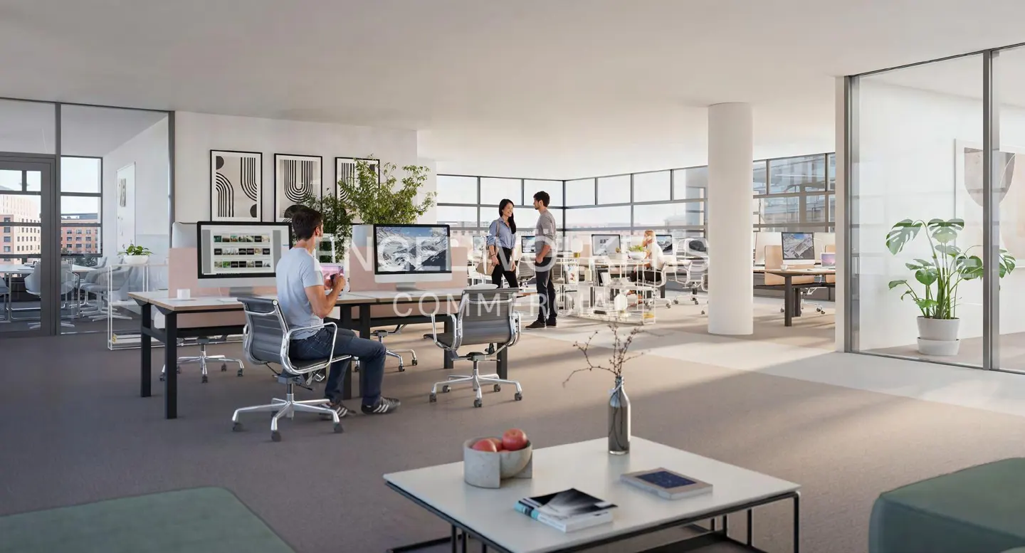 Open office space with desks, computers, and people working. Large windows provide natural light. Modern design with neutral colors.