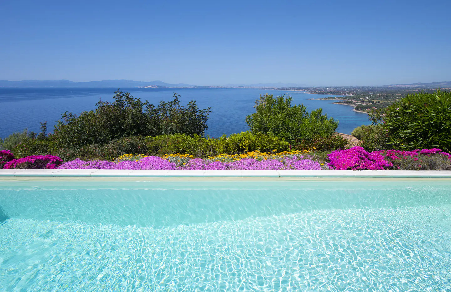 A turquoise pool overlooks the ocean under a clear blue sky. Colorful flowers and green bushes line the pool's edge.