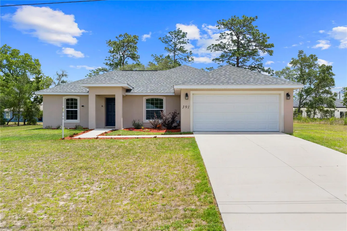 Beige single-story home with a gray roof, white garage door, and a long concrete driveway under a blue sky.