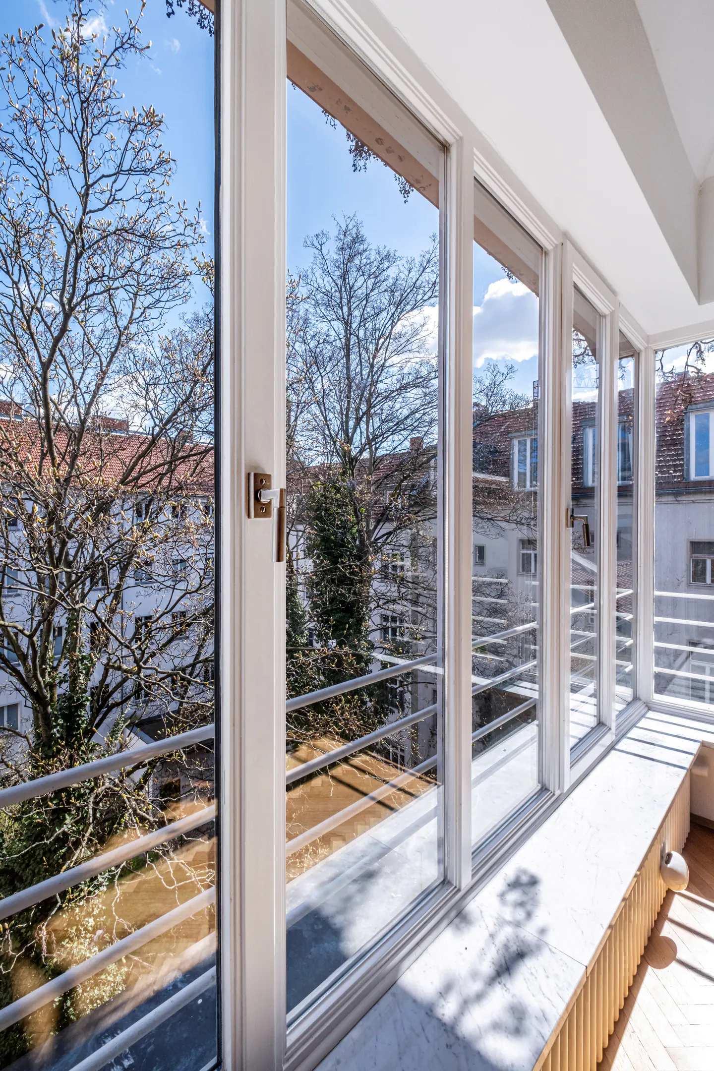 Bright room with white-framed windows overlooking a balcony with trees and buildings in the background on a sunny day.