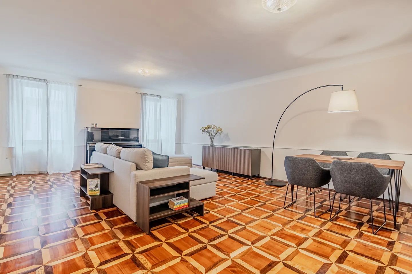 Bright living room with patterned wood floors, a beige sectional sofa, a dining table with grey chairs, and a floor lamp.