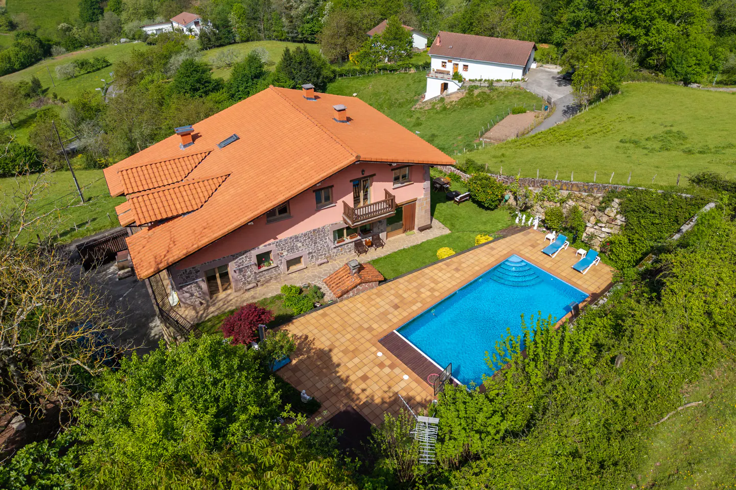Aerial view of a pink house with an orange tile roof and a blue swimming pool in a green, hilly landscape.