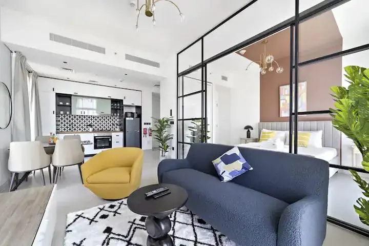 Bright studio apartment with a blue sofa, yellow chair, and black-framed glass partition separating the living area from the bedroom.
