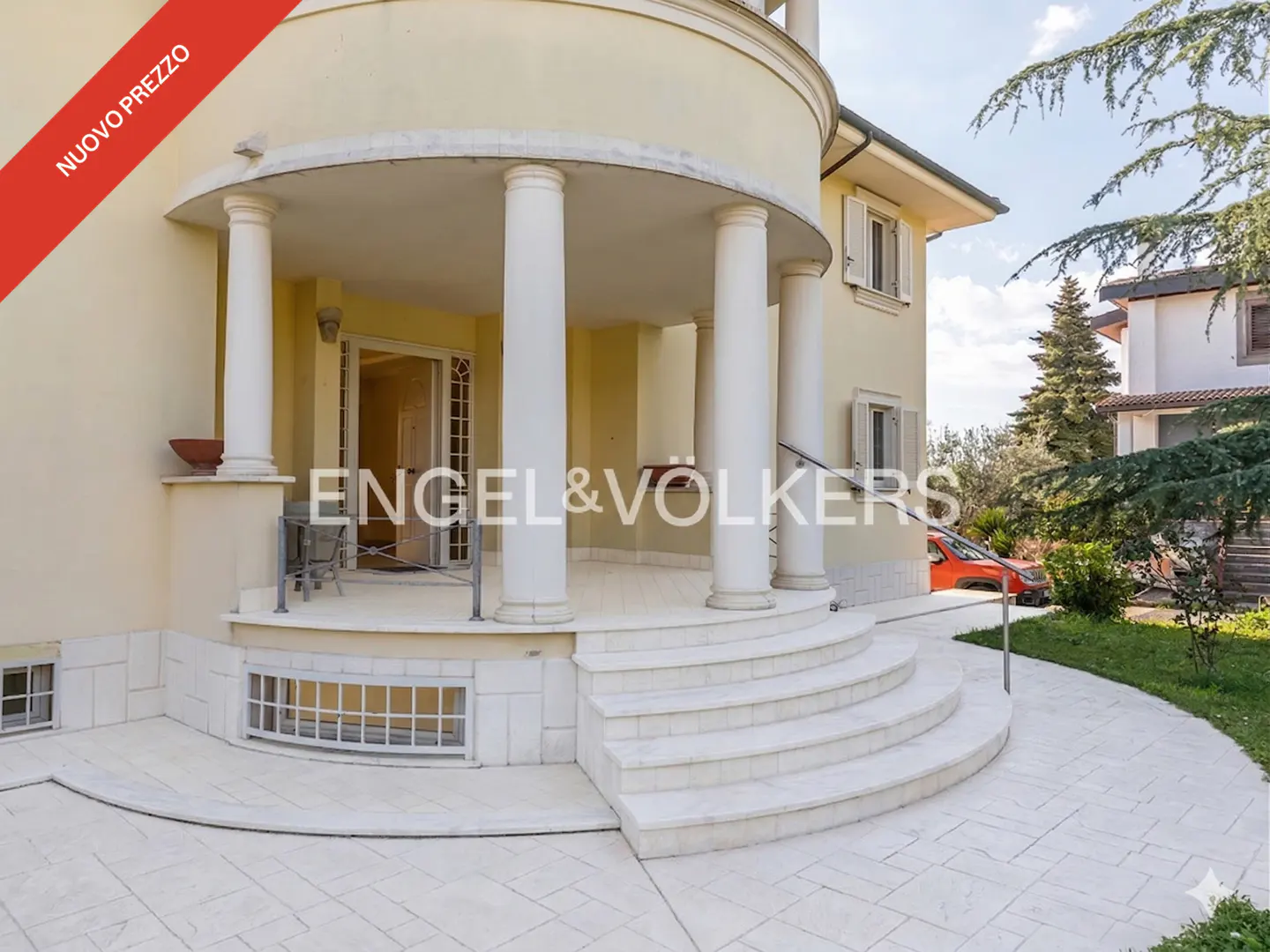 Cream-colored villa with a curved porch, white columns, and stone steps leading to the entrance, set in a garden.