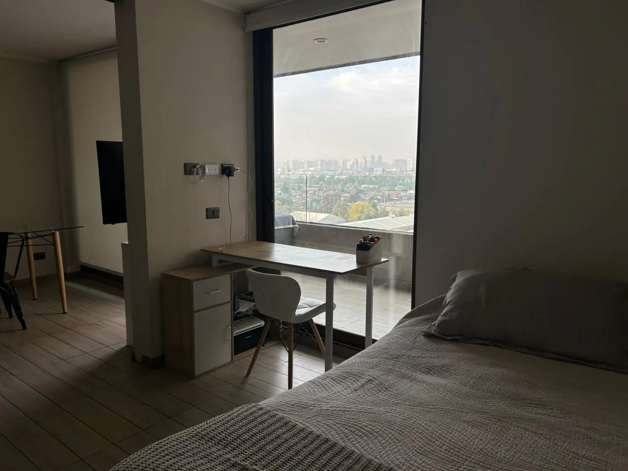 Bedroom with a desk by a large window overlooking a city skyline. Bed with gray striped blanket in the foreground.