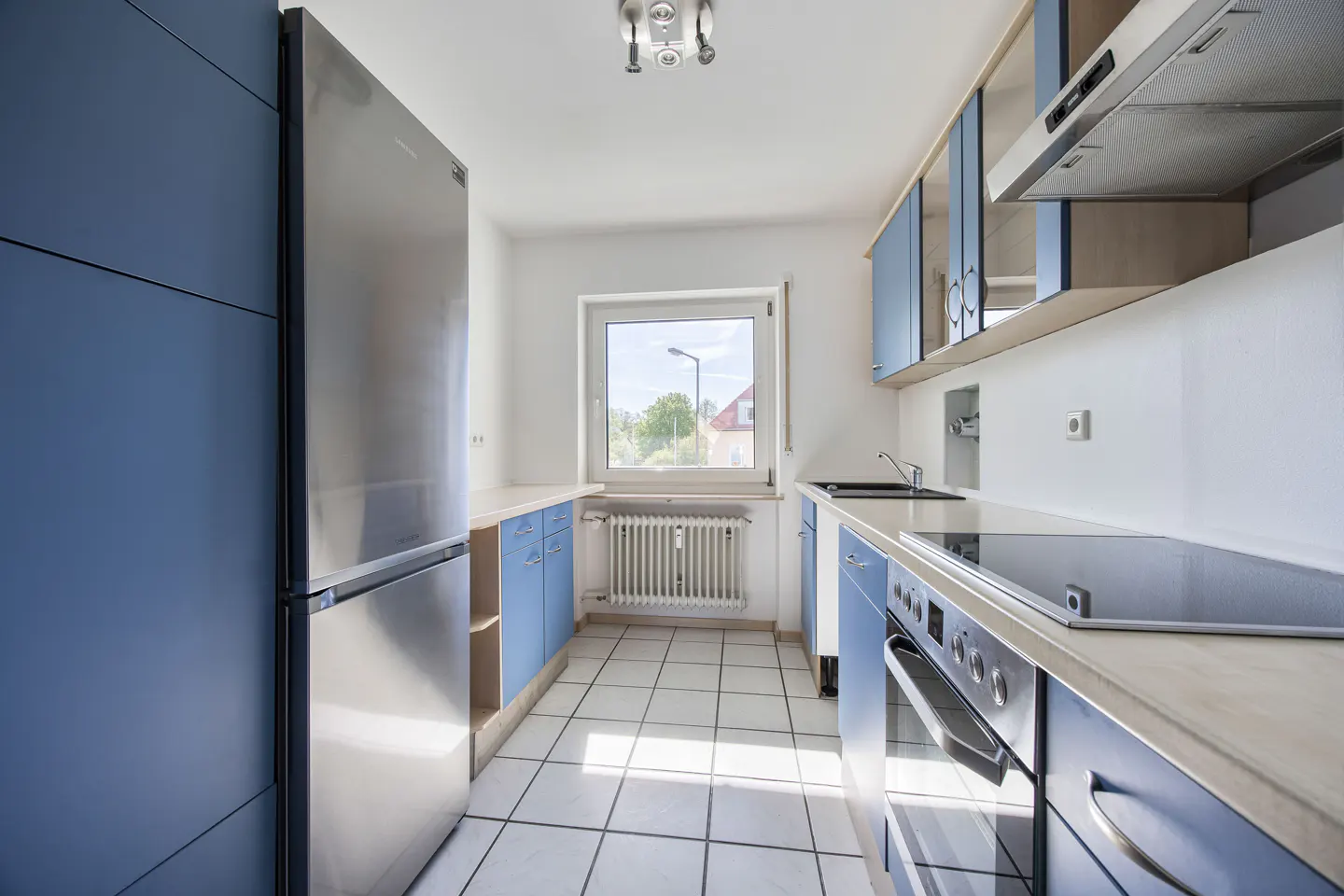 Bright kitchen with blue cabinets, stainless steel fridge, white tile floor, and window with natural light.