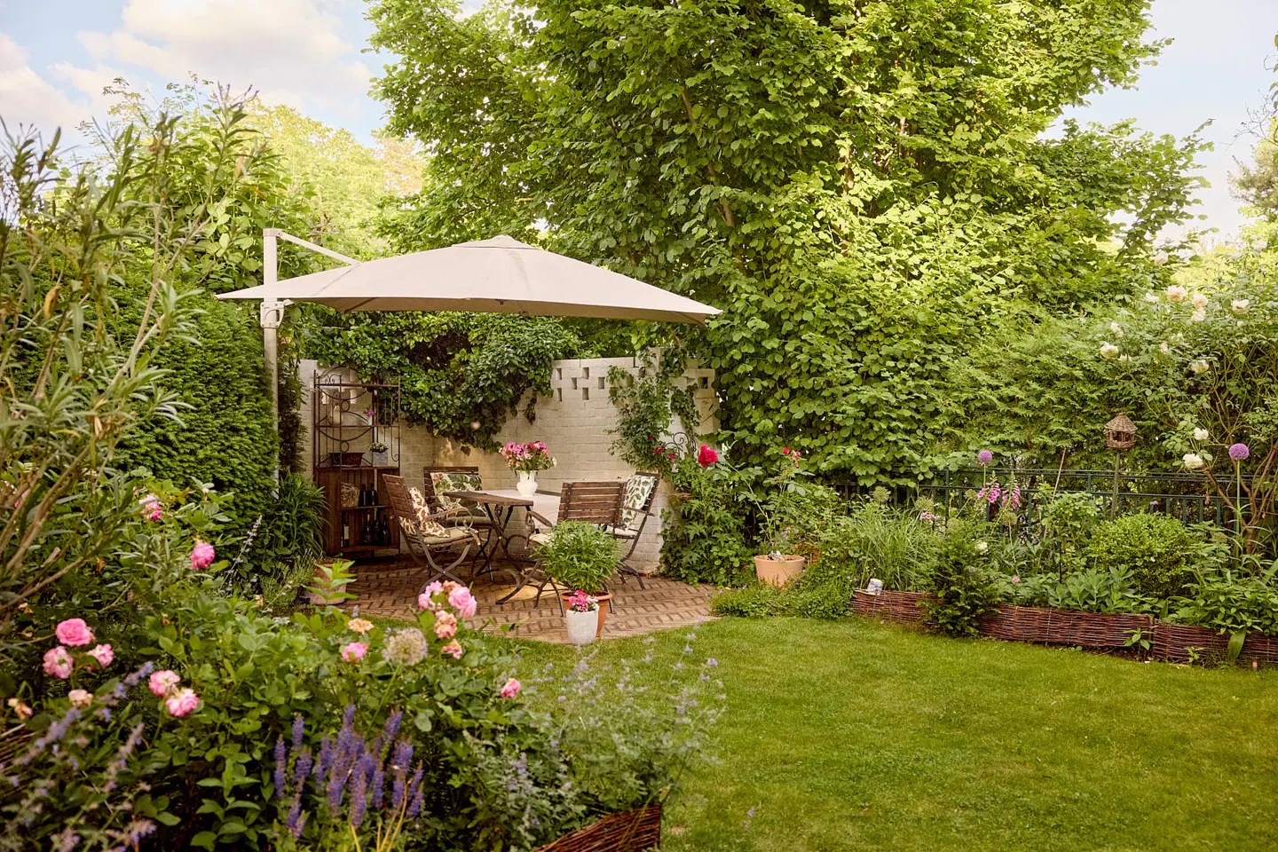 Lush garden with a patio set under a beige umbrella. Pink roses and green foliage surround a brick patio with a table and chairs.