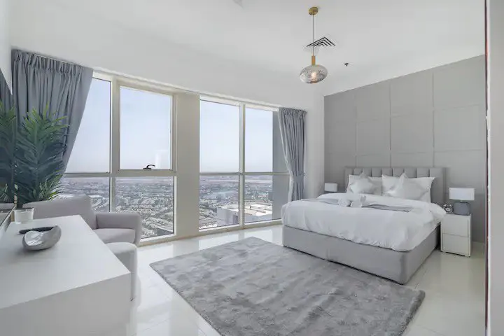 Bright bedroom with a large window overlooking a city. Gray bed, rug, and curtains. White desk and chair.
