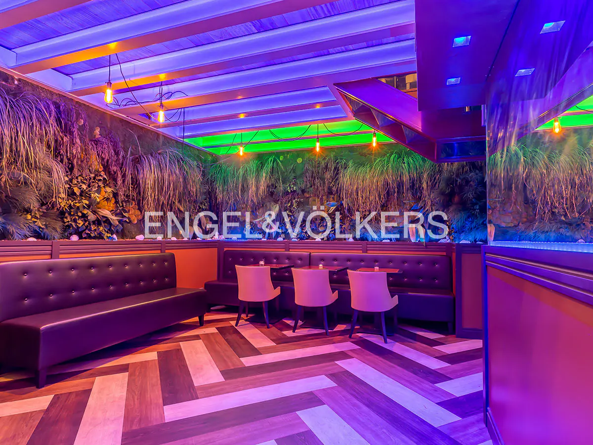 Interior view of a restaurant with purple leather booths, pink chairs, and a herringbone floor under blue and green neon lighting.