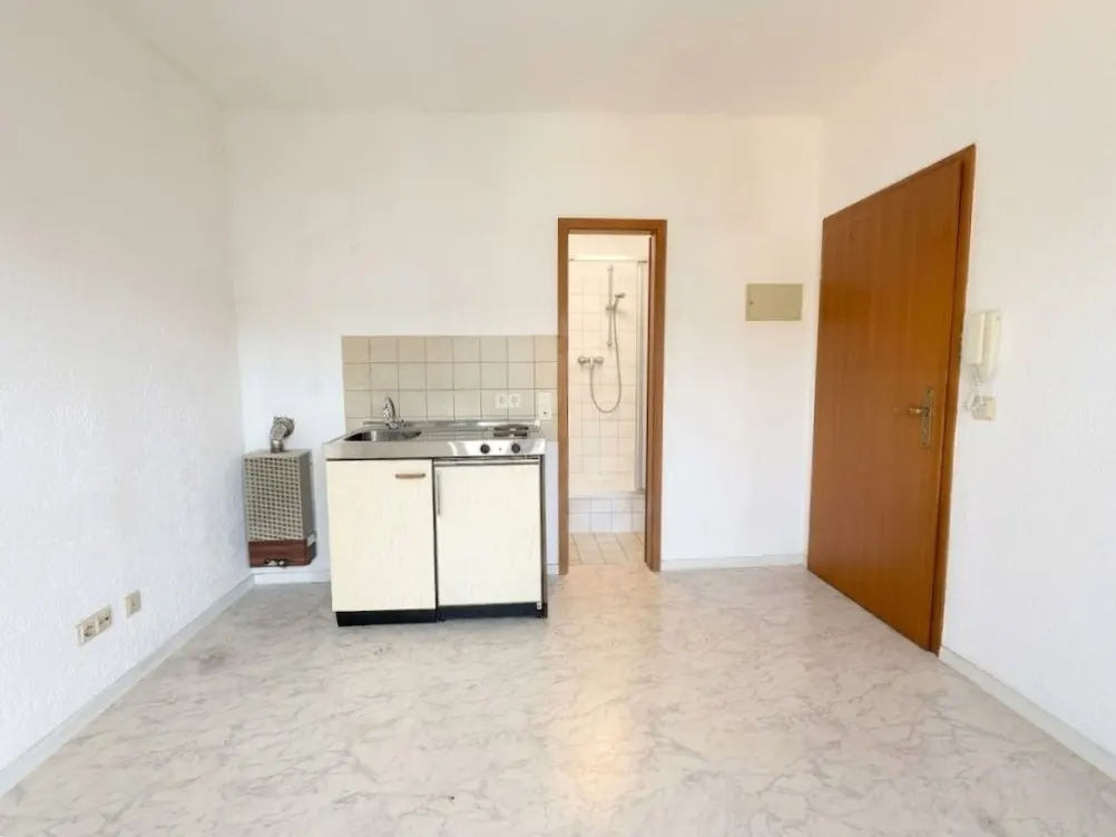 A small studio apartment with a kitchenette, a bathroom door, and a wooden door.