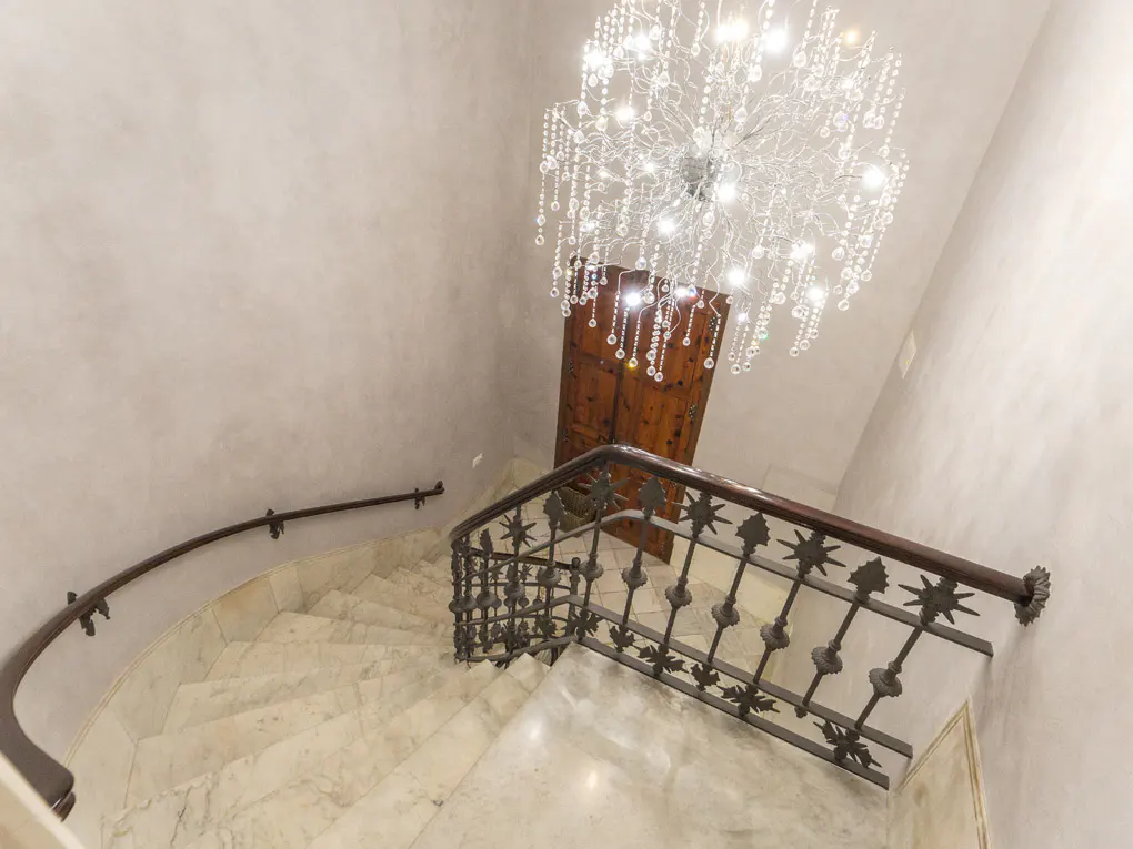 A spiral marble staircase with a dark wood and metal railing, lit by a crystal chandelier.