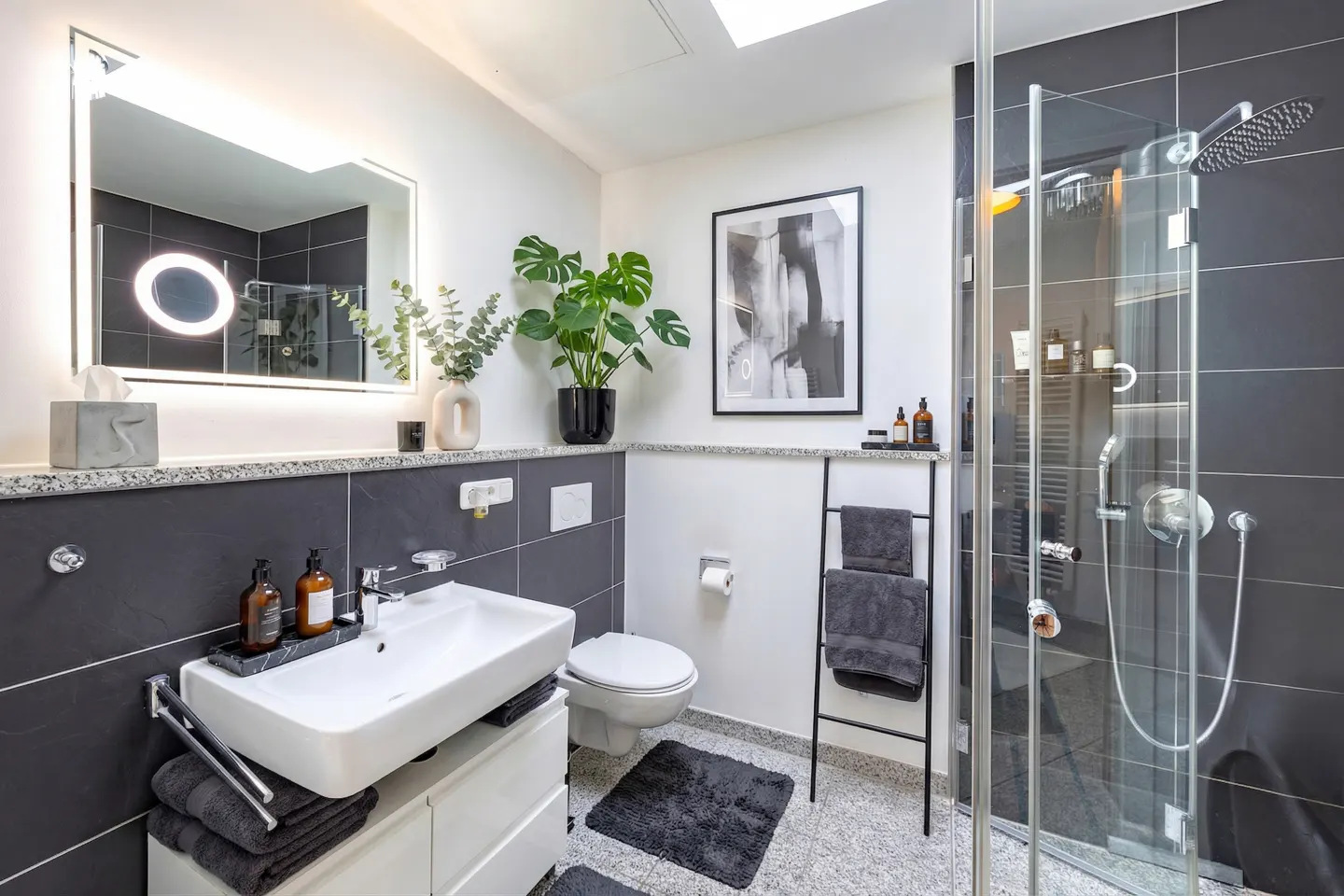 A modern bathroom with gray tile, a white sink, toilet, and shower with glass doors. A black towel ladder and plants add style.