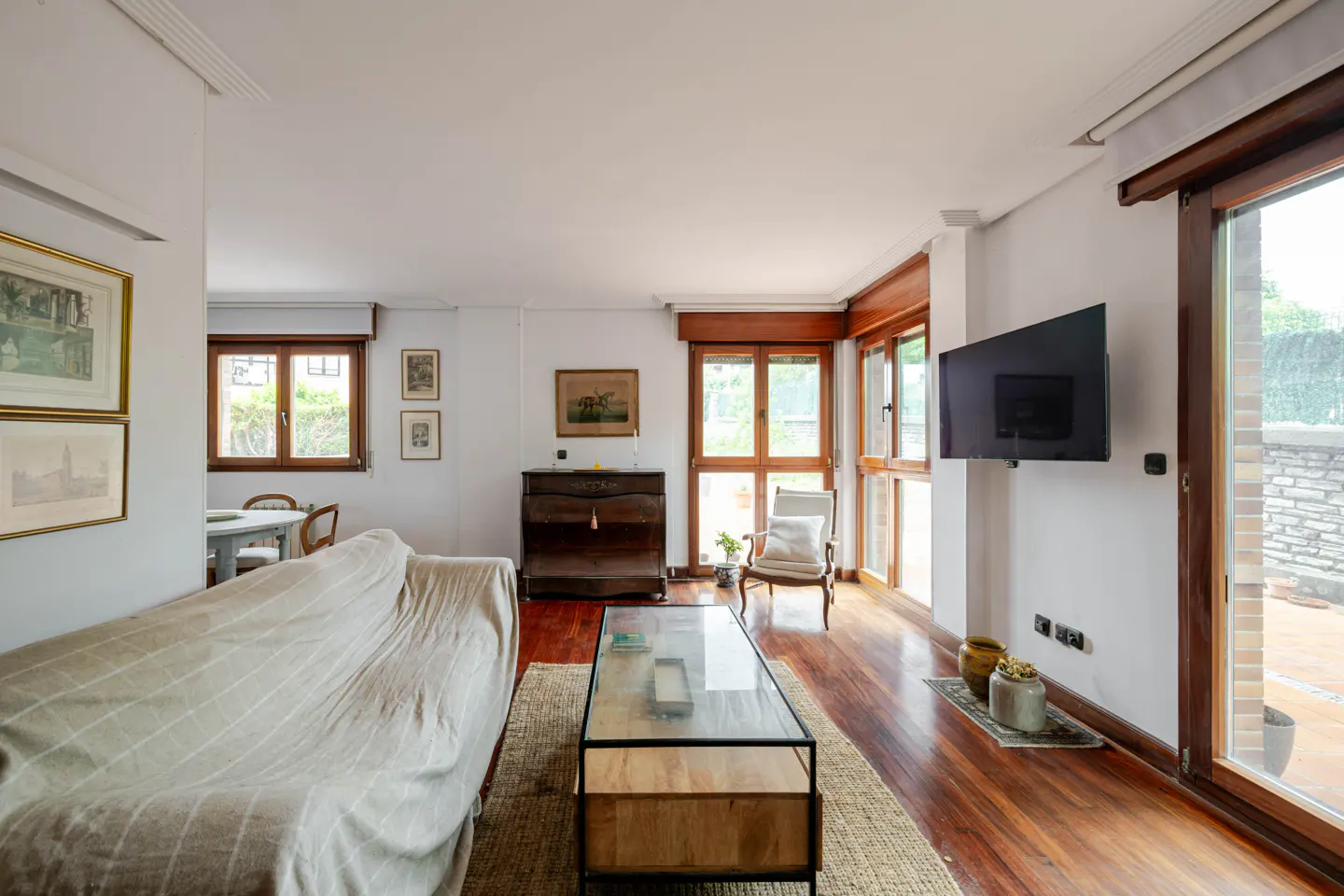 Bright living room with a beige sofa, glass coffee table, wooden floors, wall-mounted TV, and large windows to a patio.