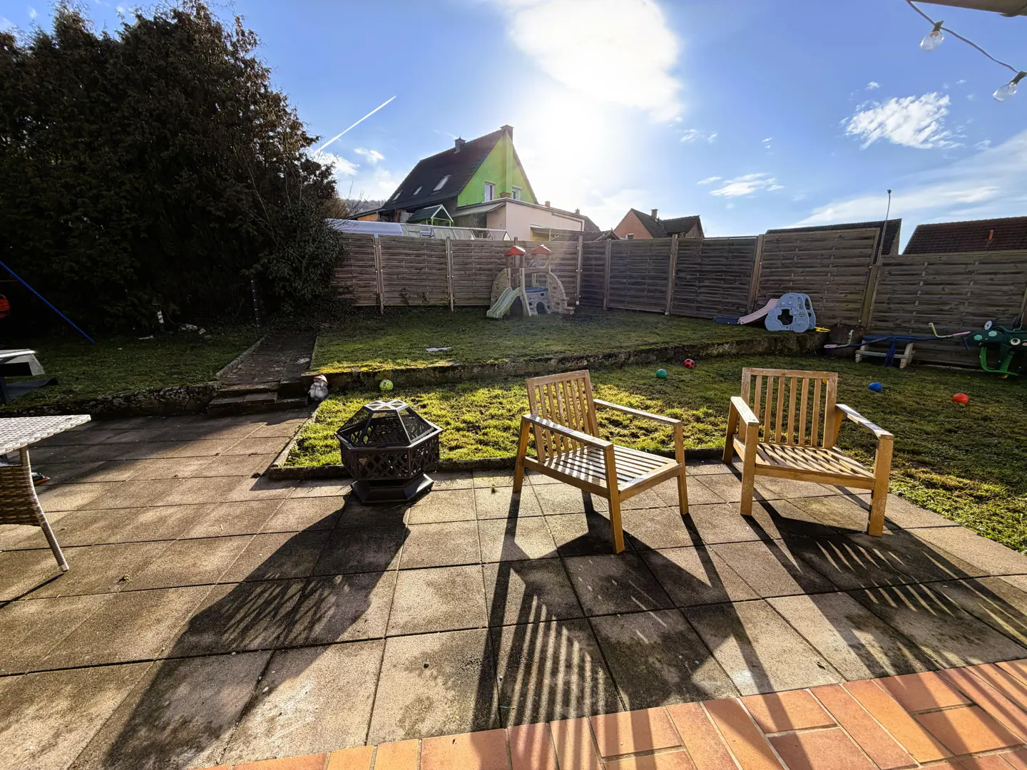 Backyard patio with two wooden chairs, a fire pit, and a grassy lawn with children's play equipment in the background.