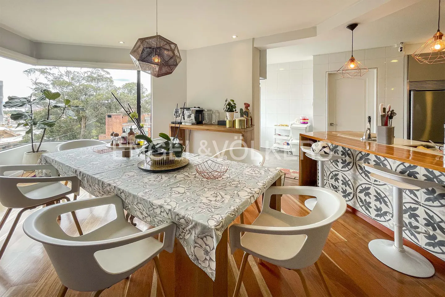 Bright dining area with a floral tablecloth, white chairs, and a geometric pendant light. A kitchen island and large window are visible.