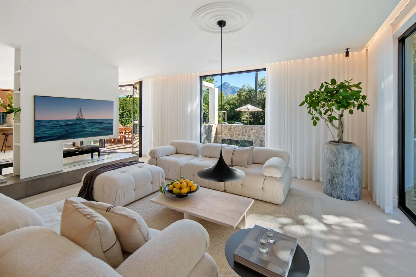 Modern living room with white modular sofas, a wooden coffee table with lemons, a large potted plant, and mountain views.