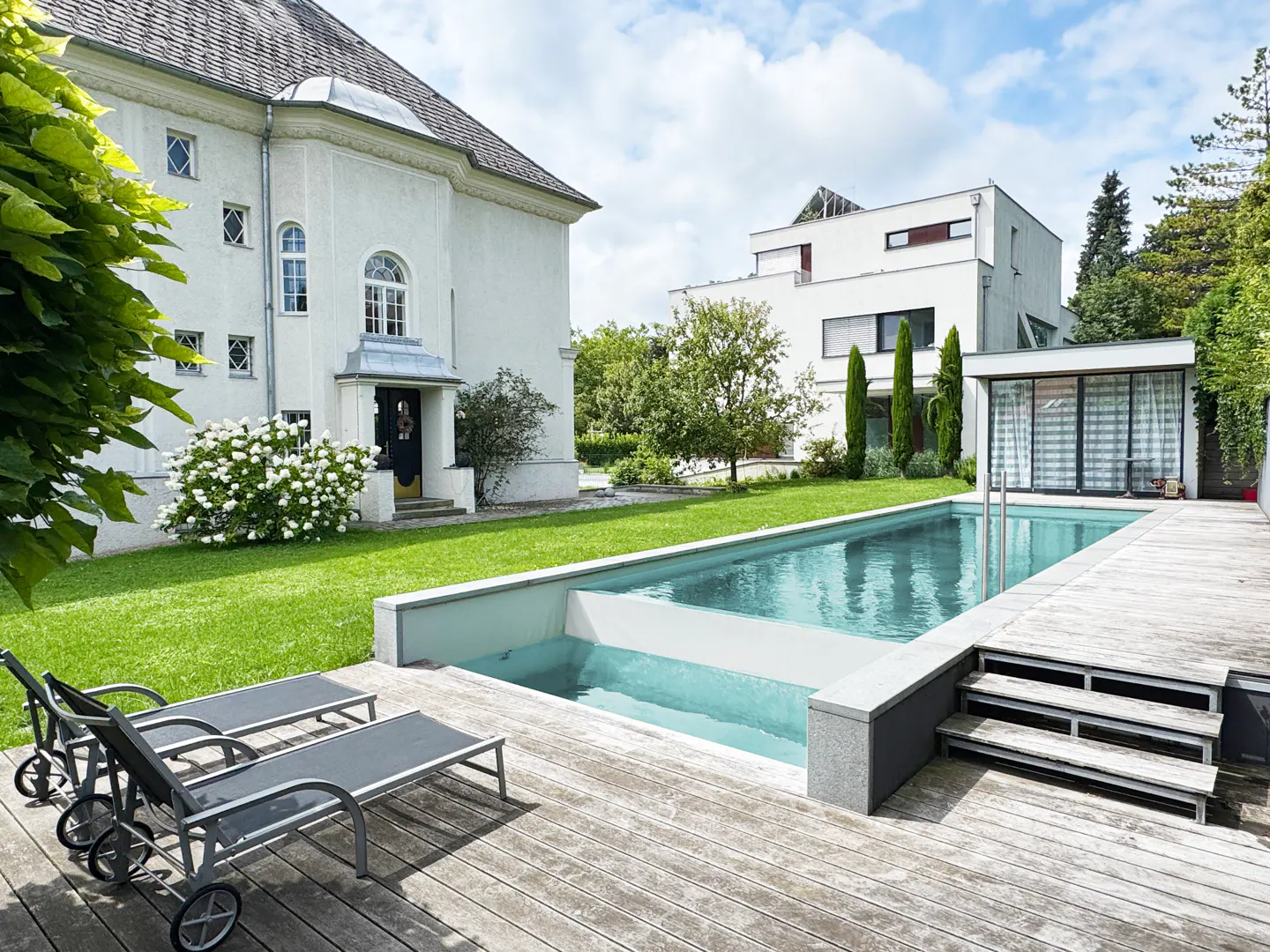 A backyard with a pool and two lounge chairs on a wooden deck, with a white house and green lawn in the background.