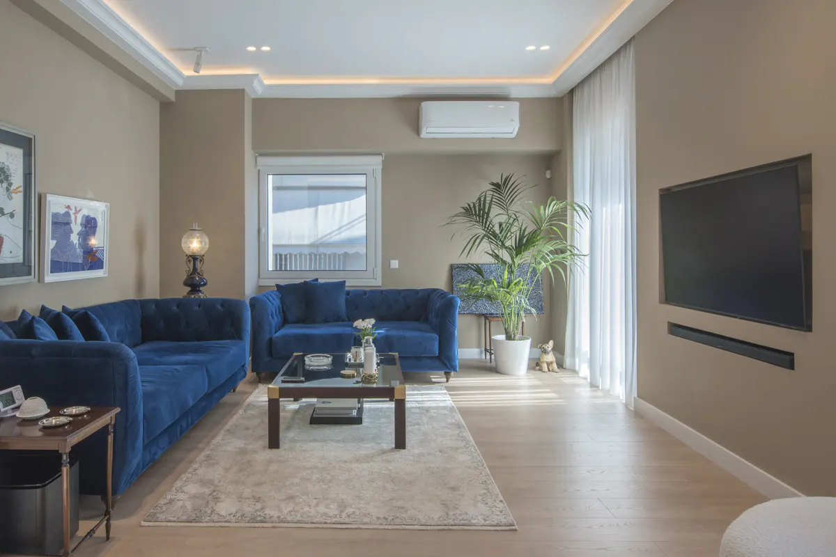 Living room with blue velvet sofas, a glass coffee table, and a large TV on a beige wall. A potted plant sits near a window with sheer white curtains.