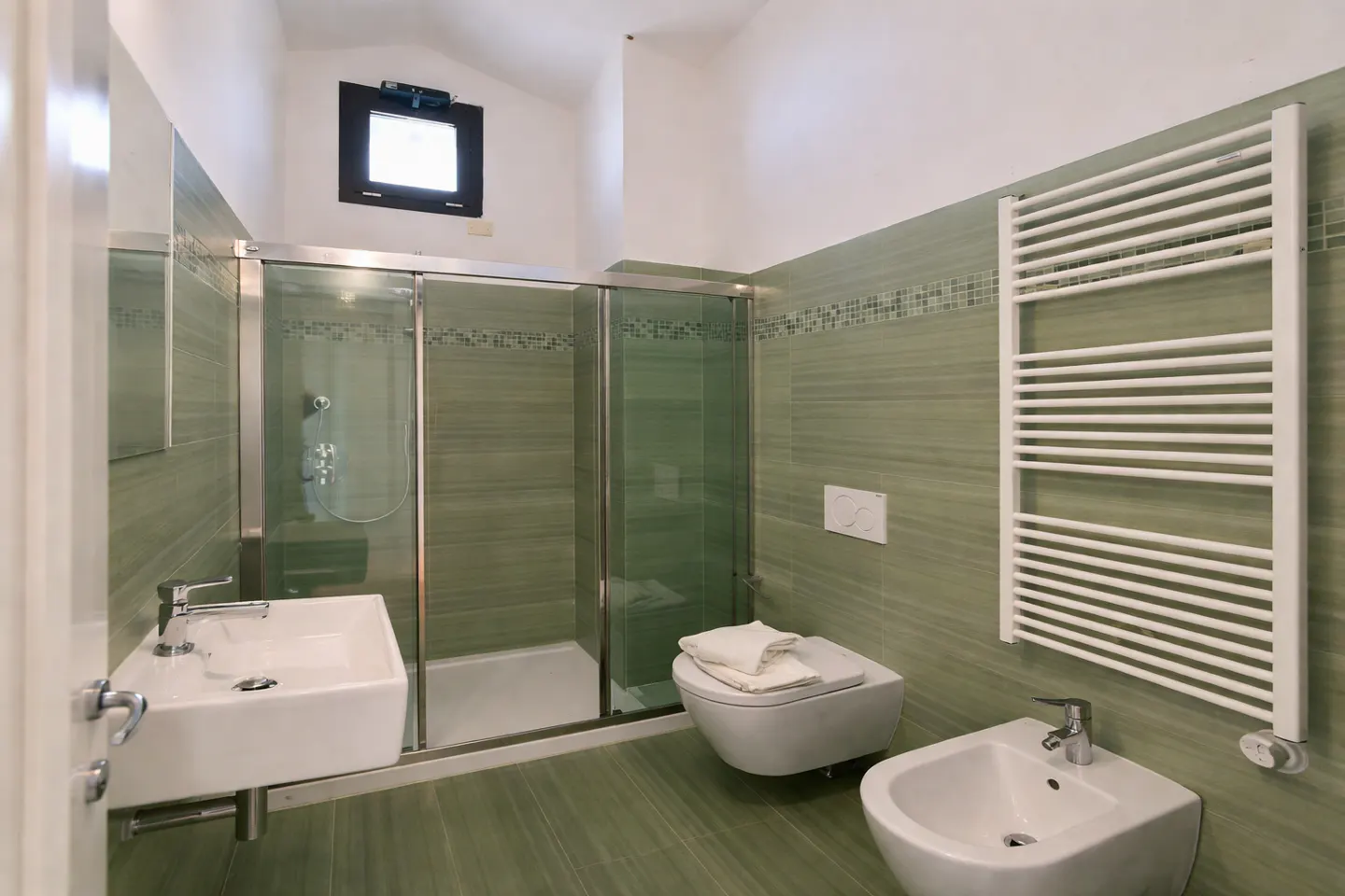 Bathroom with green tiled walls, a glass-enclosed shower, a white sink, toilet, bidet, and a white towel warmer.