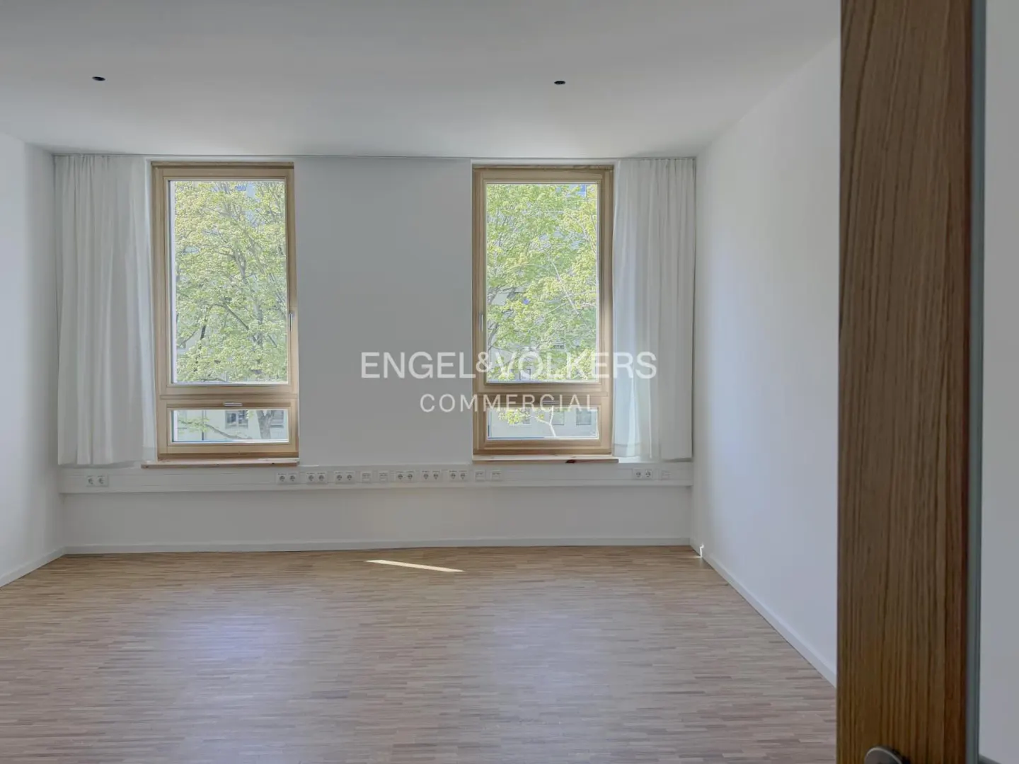 Empty office space with wood floors, white walls, and two windows with white curtains. Trees are visible outside.