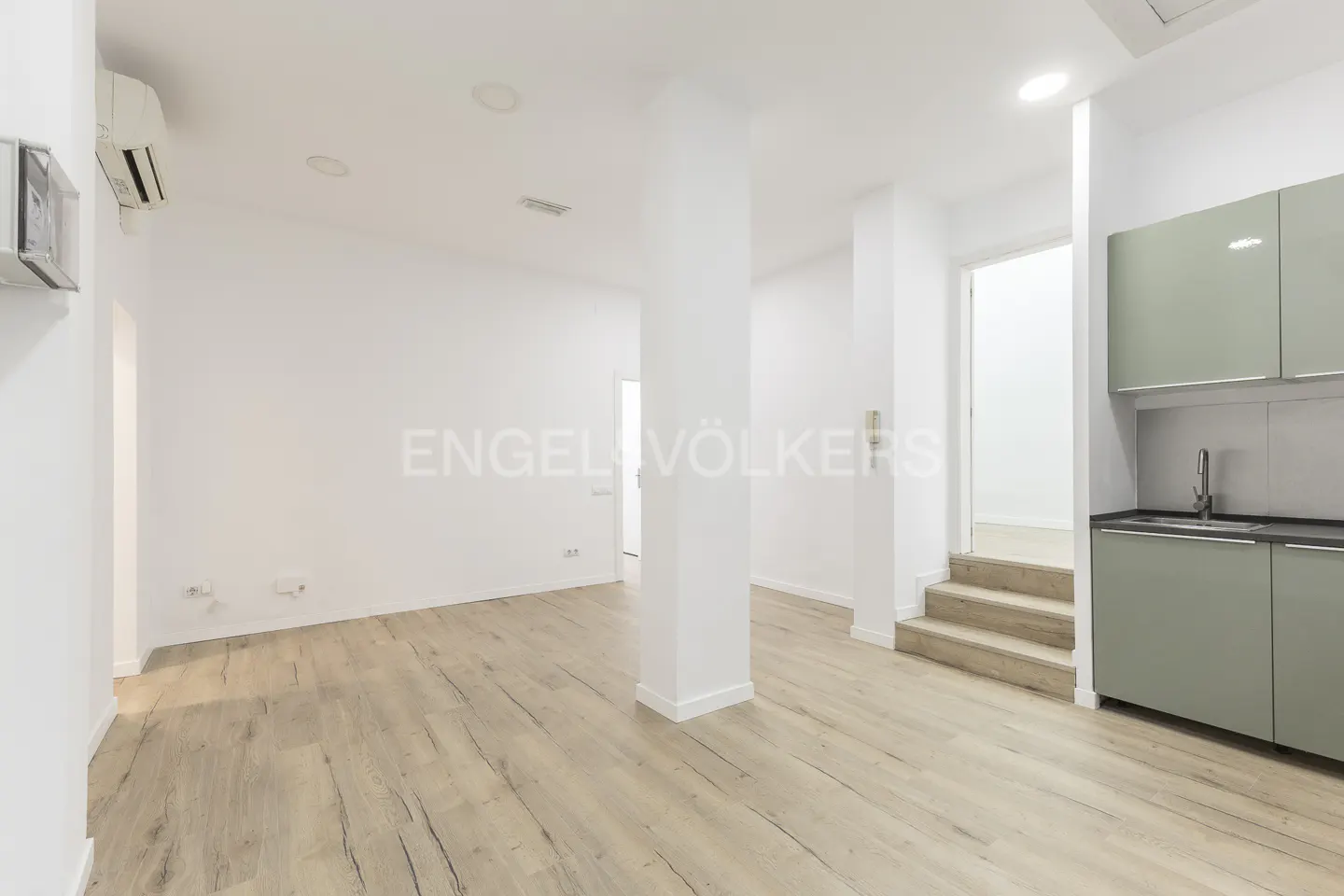 Bright, modern apartment interior with light wood floors, white walls, and sage green kitchen cabinets. A white column divides the space.