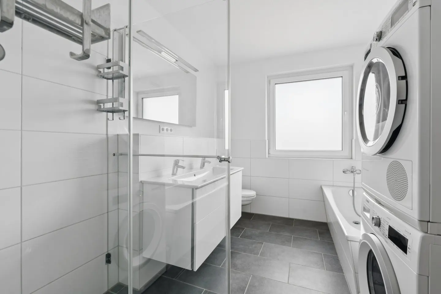 Bright bathroom with glass shower, white vanity, and stacked washer/dryer. Gray tile floor and white tiled walls.