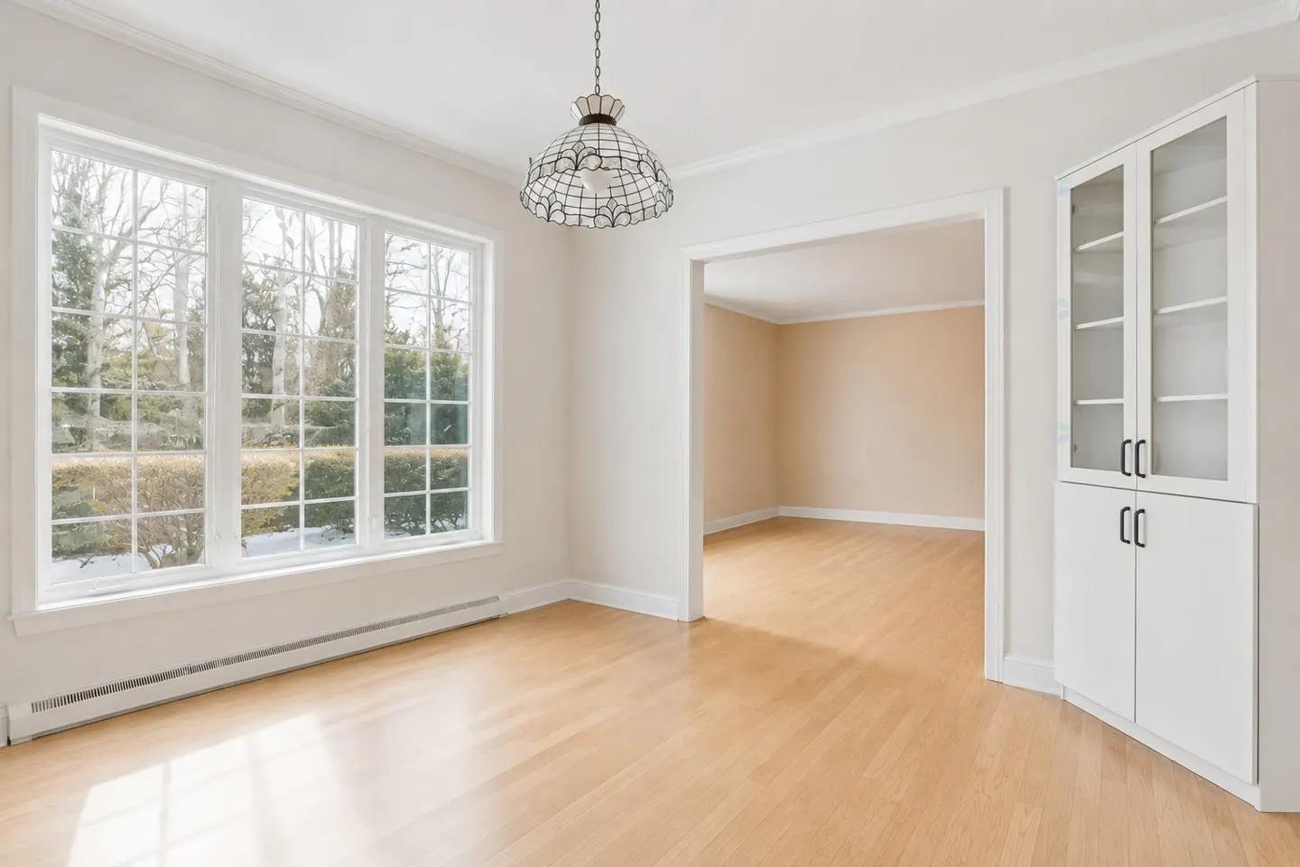 Bright, empty dining room with hardwood floors, large window, white walls, and a built-in cabinet. A doorway leads to another room.