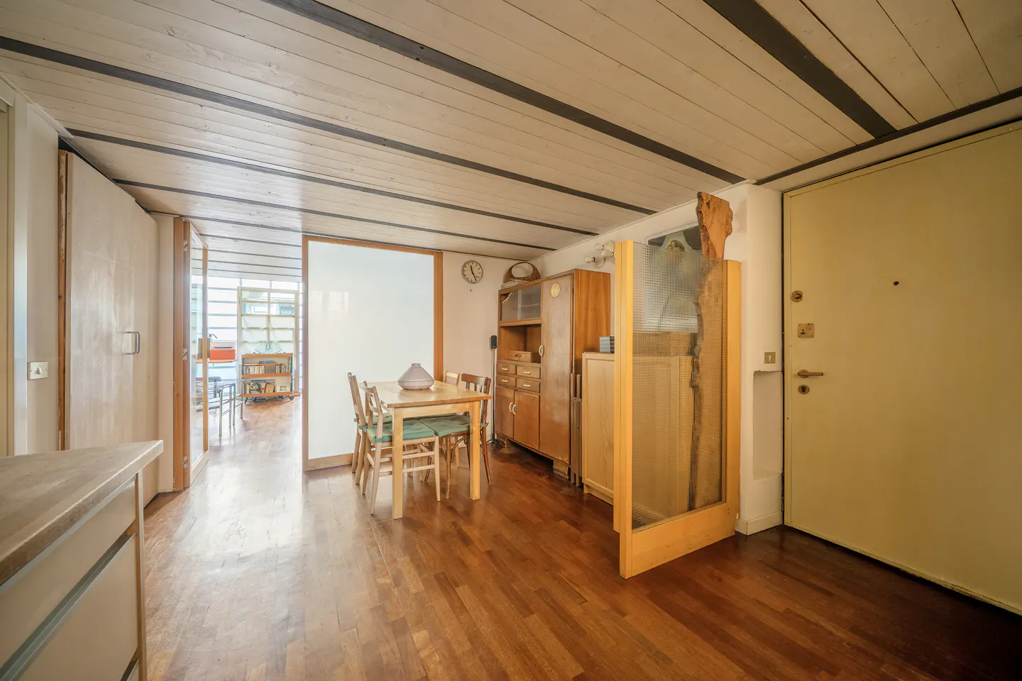 A bright, open-concept dining room with wood floors, a table with four chairs, and a vintage wooden cabinet. A white door and a glimpse into another room are visible.