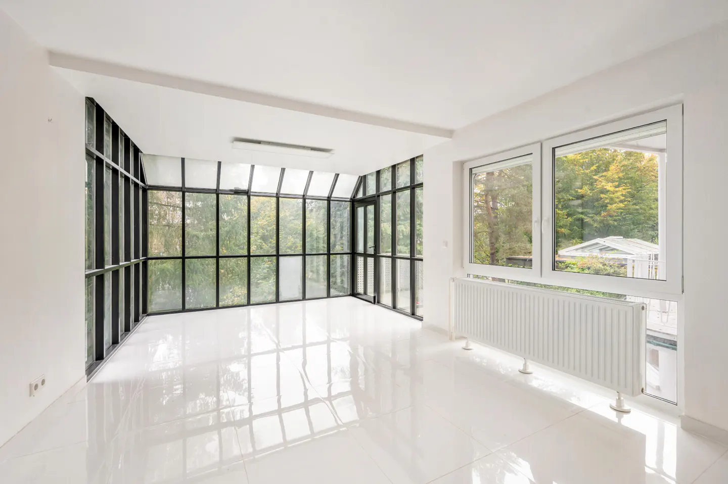 Bright, empty sunroom with white walls and tile floor. Large black-framed windows offer a view of green trees. A white radiator sits under a smaller window.