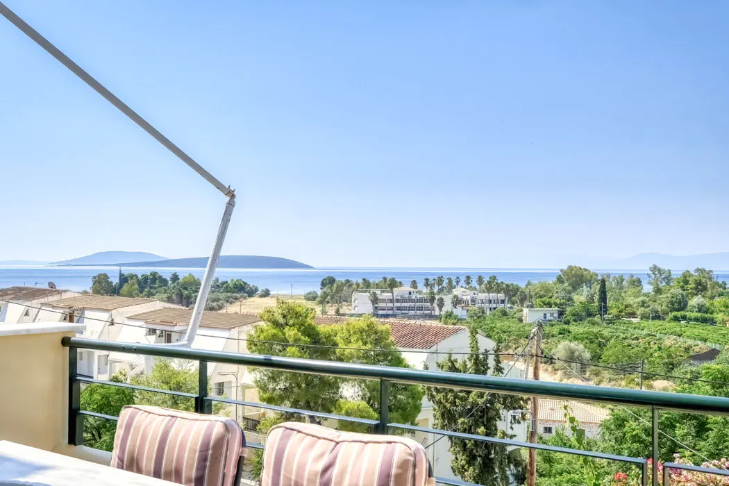 Balcony view of a coastal town with a blue sea and sky. Green trees and white buildings are visible. Two striped cushions sit on chairs.