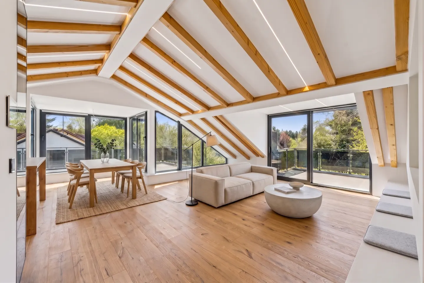Bright living room with wood floors, exposed beams, and large windows. A dining table, sofa, and round coffee table furnish the space.