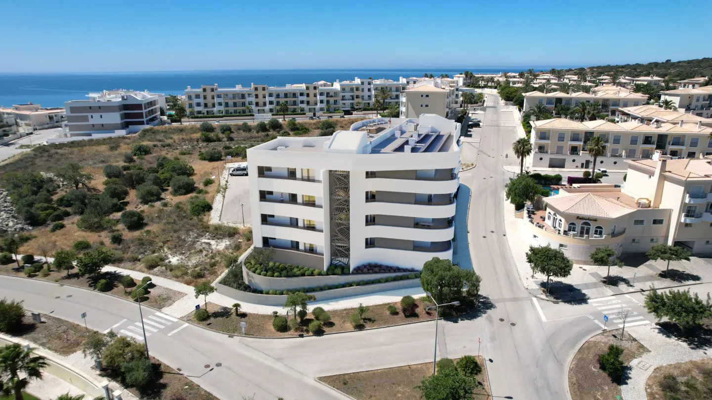 Aerial view of a modern white apartment building with curved balconies near a coastal road and the blue ocean.