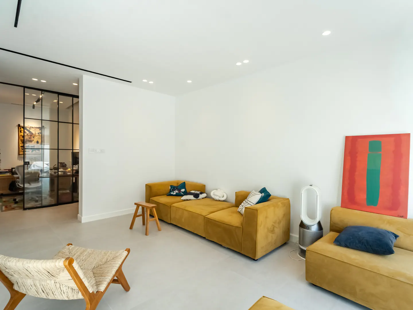 Bright living room with white walls, gray floors, and mustard-colored sofas. A glass-walled office is visible in the background. Modern art and furniture accent the space.