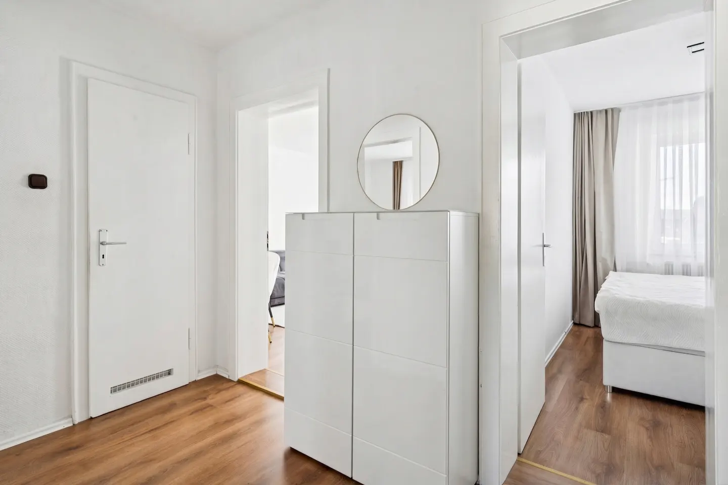 Hallway with wood floors, white walls, and doors. A white cabinet with a round mirror sits between doorways to other rooms.