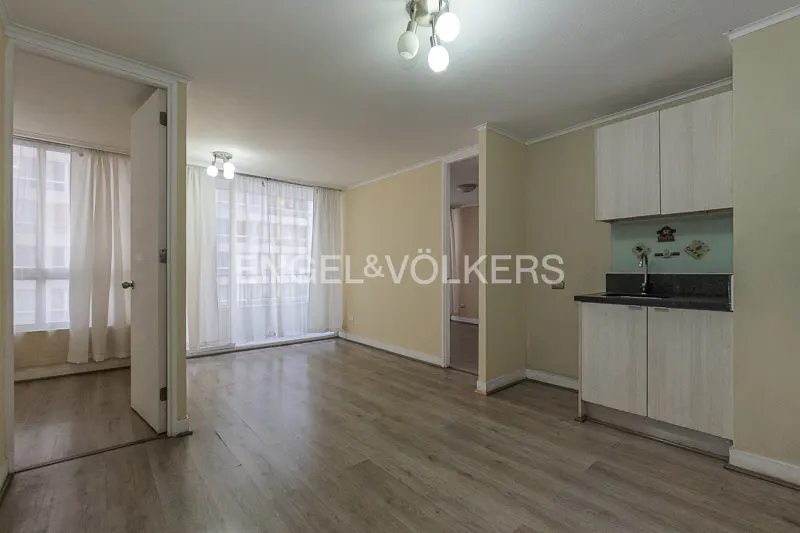 Open-plan apartment with light wood floors, beige walls, a small kitchenette, and large windows with white curtains.