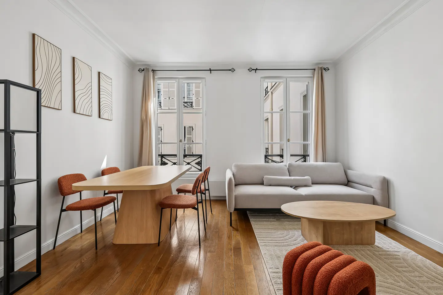 Bright living room with hardwood floors, a light gray sofa, and a light wood dining table with orange chairs. Two windows with beige curtains.
