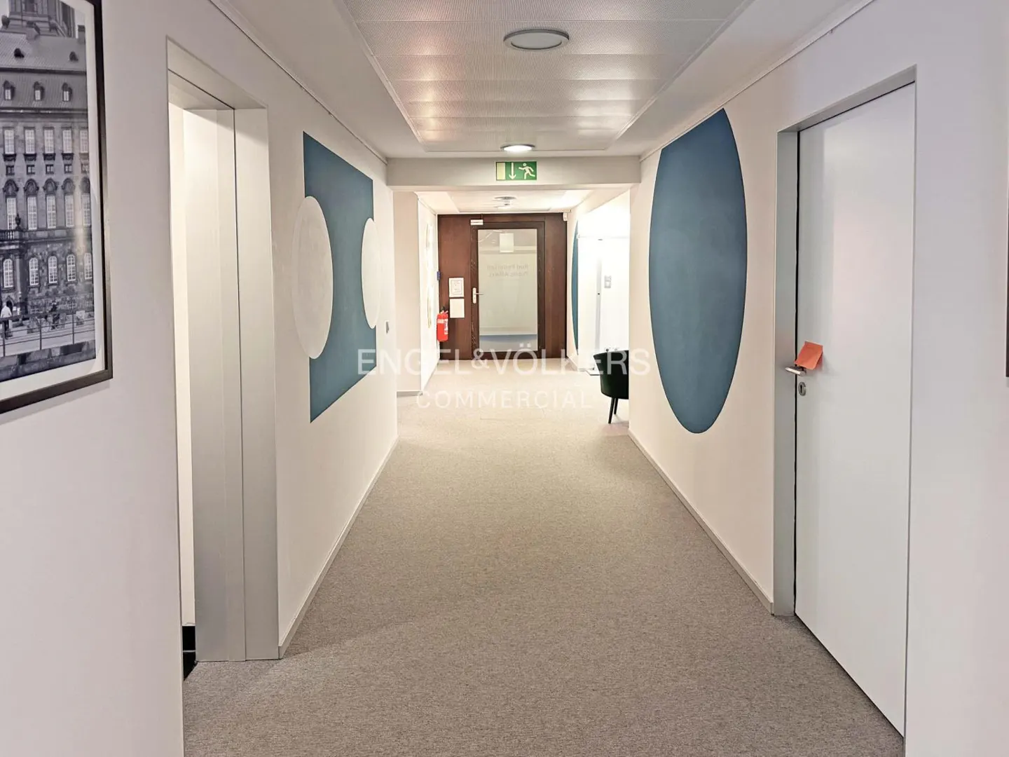 Hallway with gray carpet, white walls, and blue geometric art. White doors line the hall. An exit sign is visible at the end of the hall.