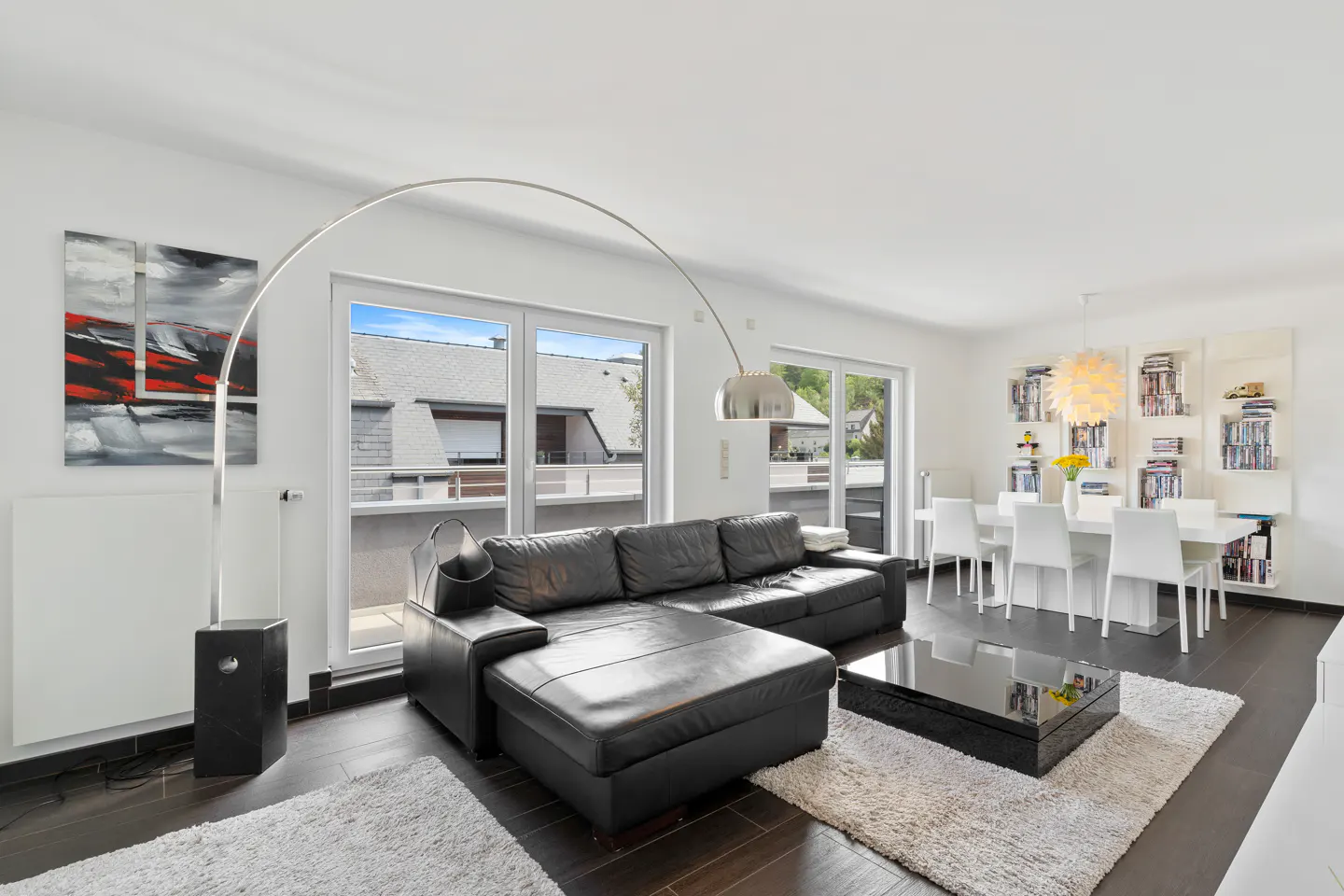 Bright living room with a black leather sofa, a modern floor lamp, and a white dining table with chairs. Abstract art hangs on the wall.