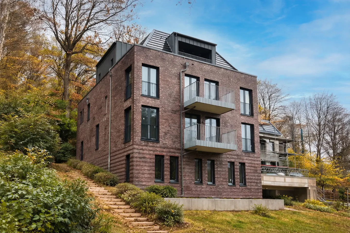 Modern brick apartment building with balconies, nestled on a hillside with trees and a stone staircase leading up to it.