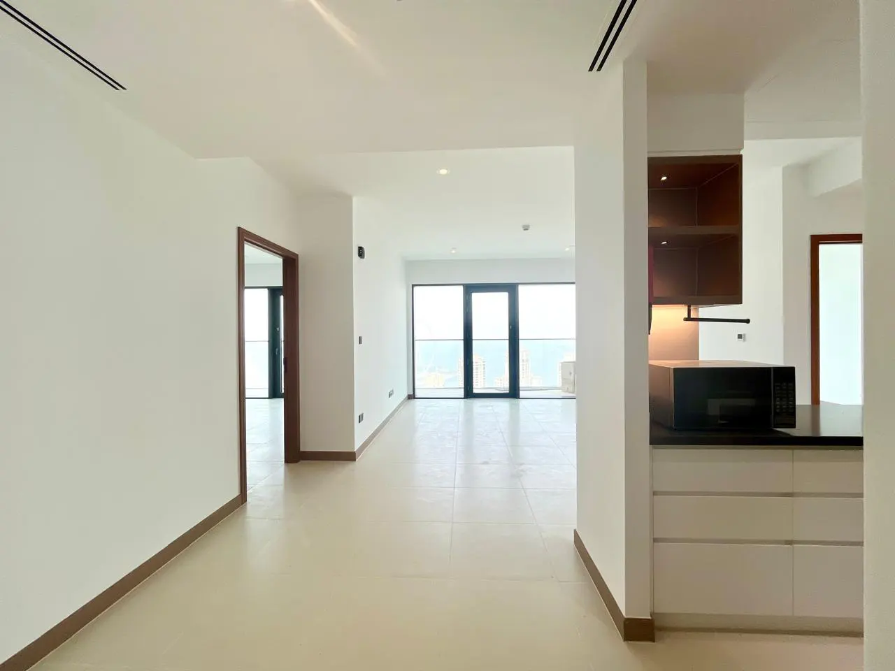 A bright, empty apartment with white walls, tile floors, and a view of the ocean through large windows. A modern kitchen is visible on the right.