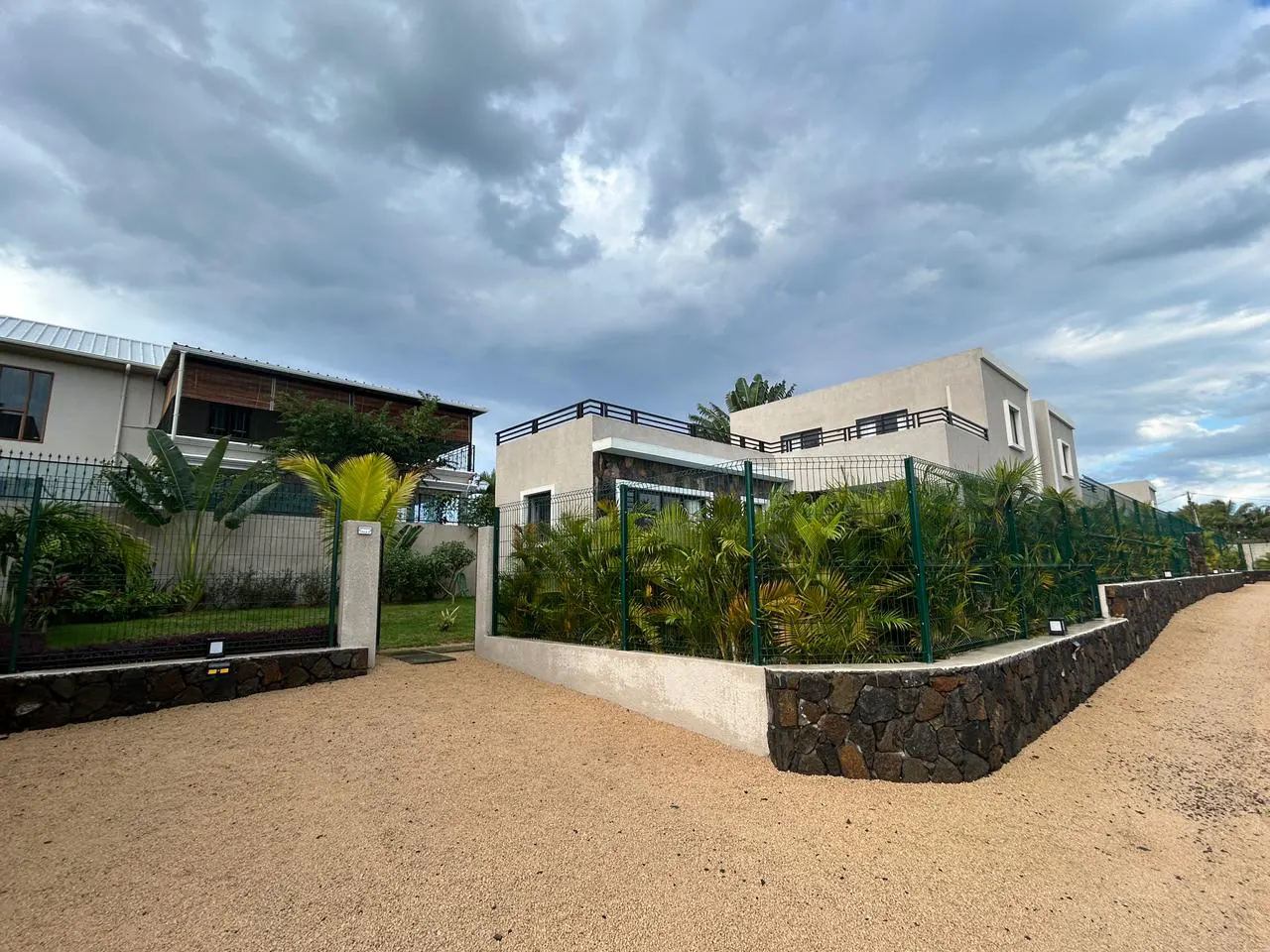 Modern beige houses with flat roofs and stone base walls behind a green fence, set against a cloudy sky.