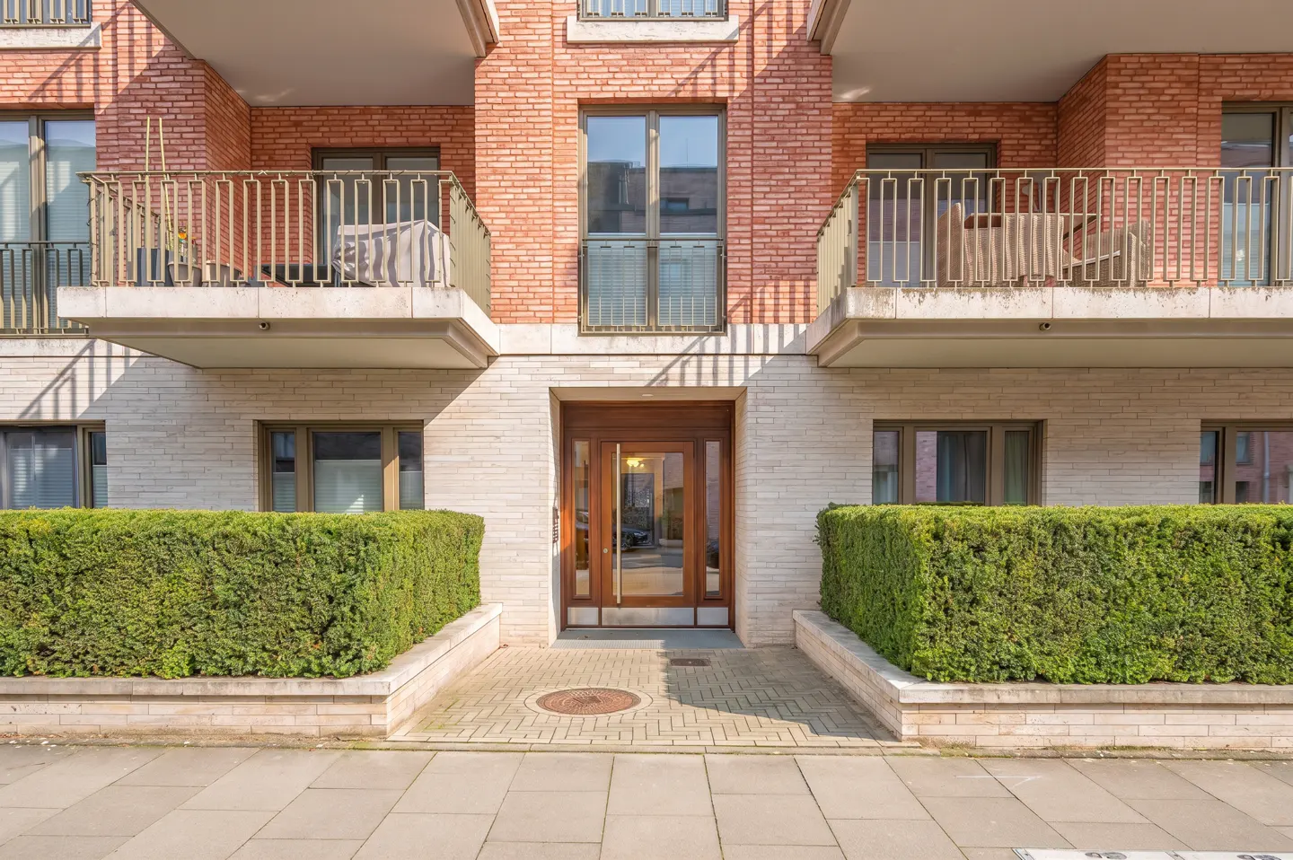 Modern apartment building entrance with red brick, light stone facade, wooden door, and manicured green hedges.