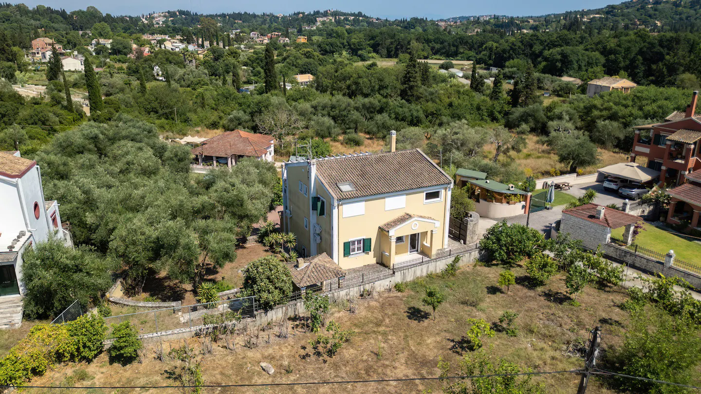 Aerial view of a two-story yellow house with a brown tile roof, green shutters, and a small porch, surrounded by trees.